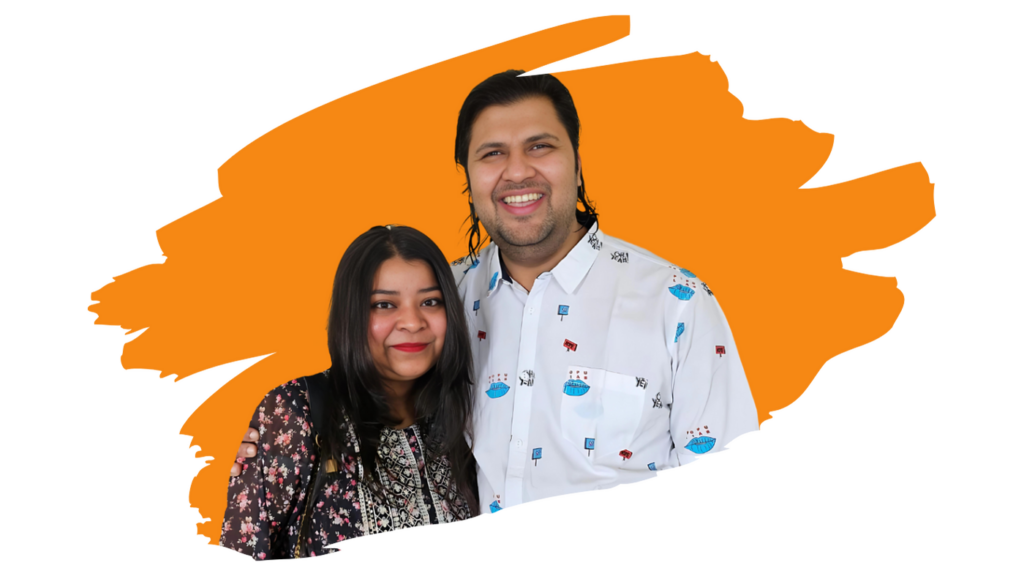 Monika and Raghavendra standing together, smiling at the camera with a bright orange abstract background. Monika is wearing a black floral-patterned top, while Raghavendra is dressed in a white shirt with various small, colorful patterns.
